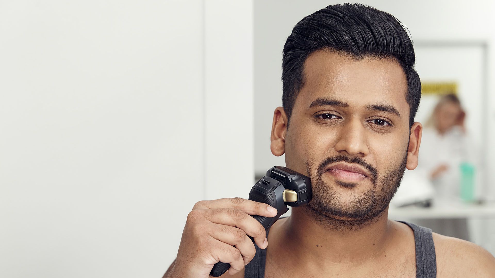 A person using an electric trimmer to clean the neckline under the jaw, head held level, with a softly curved neckline visible in the mirror